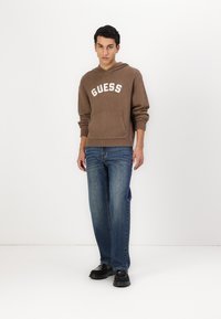Bruine hoodie met 'GUESS' logo, voorzak, gecombineerd met blauwe rechte jeans en zwarte schoenen, tegen een effen witte achtergrond.