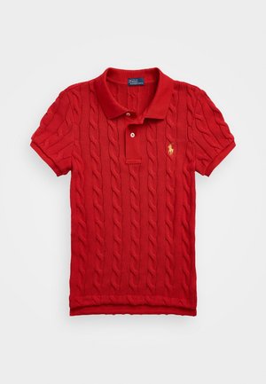 Punainen kabelineuleinen polo-paita, jossa on kaulus, kolmen napin lista ja kultaisella brodeerattu logo. Pehmeä textura ja lyhyet hihat.