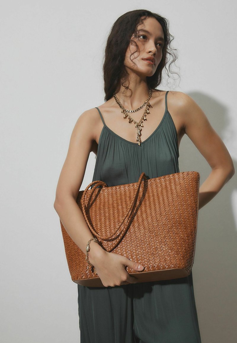 Femme en robe sans manches vert foncé tenant un grand sac cabas en osier marron, portant un collier doré superposé et un bracelet, se tenant devant un fond uni.