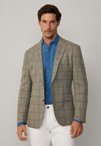 Blazer a pied-de-poule in tessuto grigio testurizzato con accenti verdi e blu, abbinato a una camicia di denim blu e pantaloni bianchi.