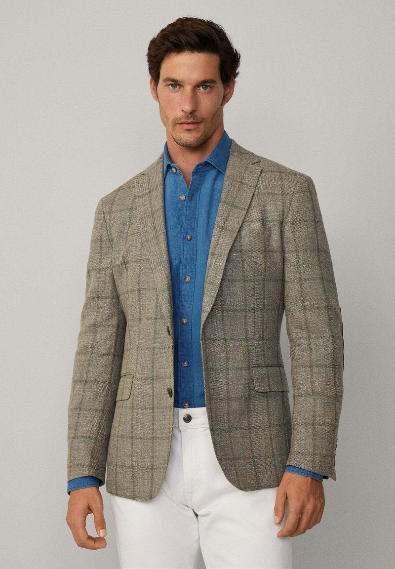 Blazer a pied-de-poule in tessuto grigio testurizzato con accenti verdi e blu, abbinato a una camicia di denim blu e pantaloni bianchi.