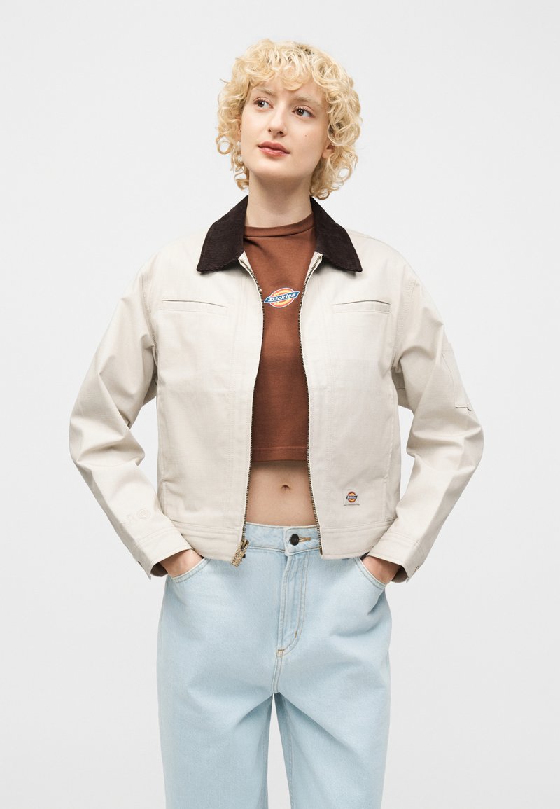 Junge Person mit kurzen, lockigen blonden Haaren, die eine beigen Jacke mit schwarzem Kragen, ein braunes kurz geschnittenes Shirt und hellblaue hoch taillierte Jeans trägt.