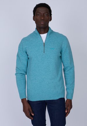 Pull turquoise à fermeture éclair en tissu tricoté texturé, doté d'un col haut et de poignets côtelés. Porté par-dessus un t-shirt blanc.