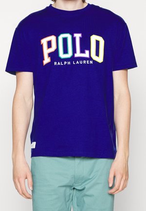 Marineblauw T-shirt met grote kleurrijke "POLO"-letters en kleinere "Ralph Lauren" tekst, gedragen door een persoon die staat tegen een effen achtergrond.