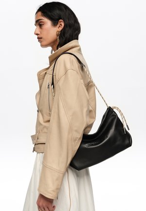 Mujer con chaqueta beige y falda blanca llevando un bolso de hombro de cuero negro con cierre de cordón, mirando hacia la izquierda sobre un fondo blanco.