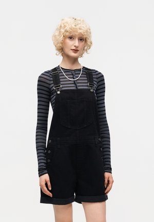 Jong persoon met krullend blond haar, gekleed in een zwarte korte tuinbroek en een grijs-zwart gestreept shirt met lange mouwen, staand tegen een effen achtergrond.