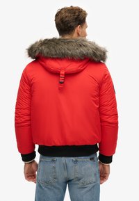 Chaqueta acolchada roja con capucha forrada de pelo, puños elásticos y un dobladillo corto. Tejido texturizado con costuras visibles y un detalle de tira en la parte posterior.