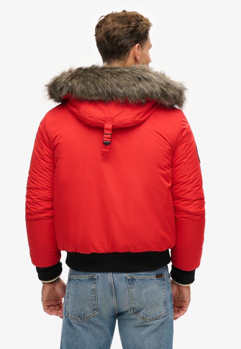 Doudoune rouge avec capuche en fourrure, poignets élastiques et ourlet court. Tissu texturé avec des coutures visibles et un détail de bandeau à l'arrière.