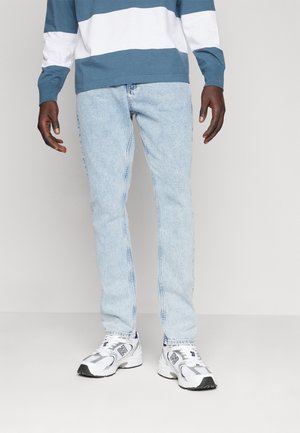 Homme portant un jean bleu clair, des baskets blanches et grises, et une chemise à manches longues rayée bleue et blanche sur un fond uni.