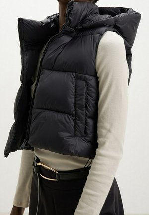Personne portant une doudoune noire avec un col montant par-dessus un haut beige à manches longues, associée à un pantalon noir et une ceinture noire avec une boucle dorée.