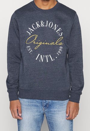 Mężczyzna w ciemnoszarej bluzie z białym i żółtym okrągłym napisem „Jack & Jones Originals Est. Intl. 1990” oraz w jasnoniebieskich dżinsach.