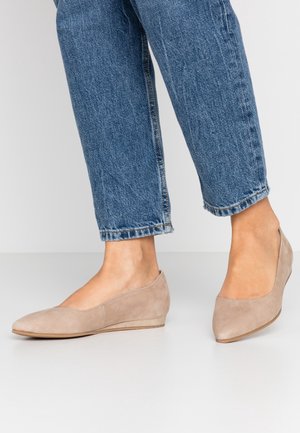 Kvinde iført blå jeans og beige ruskind lave kilehælssko står på en hvid gulv med en ensfarvet baggrund.