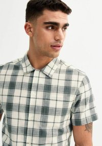 Camicia a maniche corte con motivo a quadri nei colori verde, grigio e bianco. Realizzata in tessuto leggero con chiusura a bottoni e colletto.