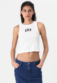 White ribbed crop top with black "GAP" logo, round neckline, and sleeveless design, paired with a dark blue denim skirt.