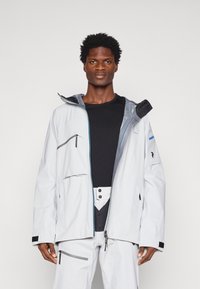 Chaqueta blanca impermeable con cremallera frontal, acentos grises, múltiples bolsillos y capucha ajustable. Combinada con una camiseta negra y pantalones grises.