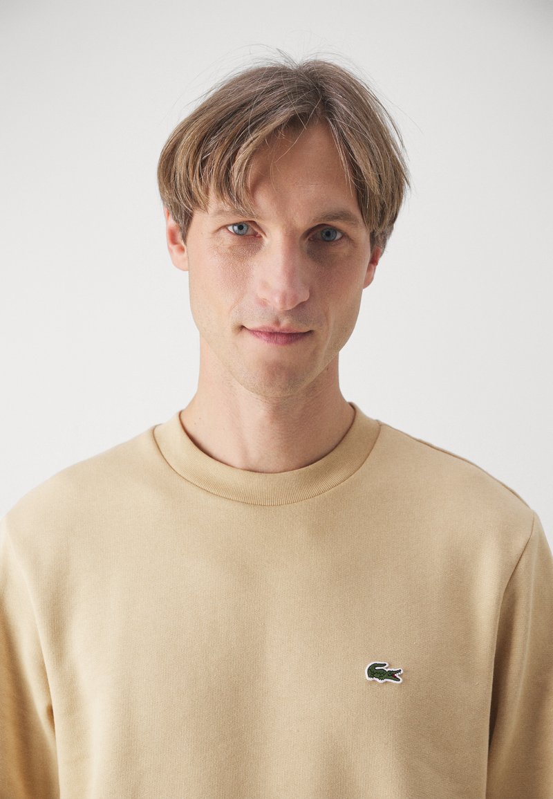 Beige Sweatshirt aus weichem Stoff, mit Rundhalsausschnitt und einem kleinen aufgestickten grünen Krokodil-Logo auf der linken Brust.