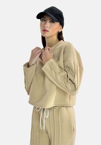 Sweat-shirt court beige à manches longues, avec des accents cousus et un col arrondi, porté avec un pantalon de jogging beige et une casquette noire.