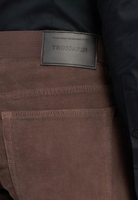 Hnědé kordových kalhoty s texturovaným povrchem a koženou nášivkou na pásku, s názvem značky "TRUSSARDI."