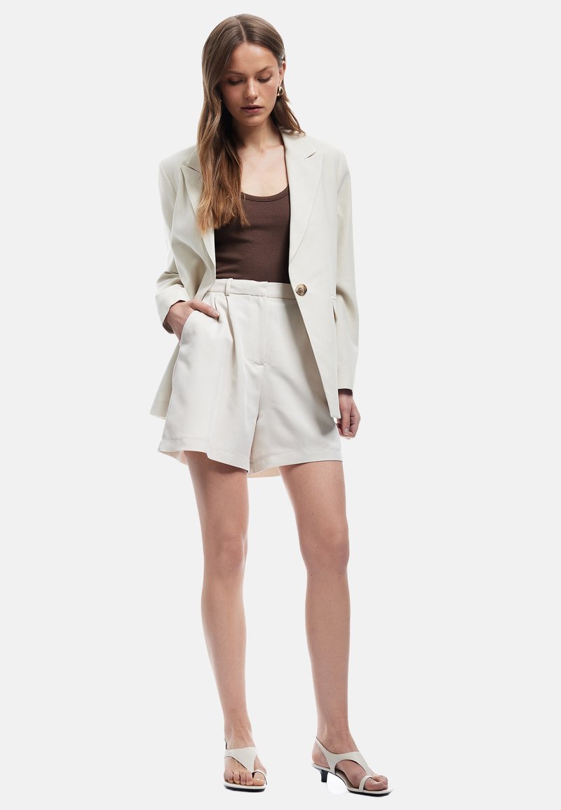 Blazer crema con un solo botón dorado, combinado con pantalones cortos blancos de talle alto. Top ajustado marrón, sandalias de tacón blancas con tiras.
