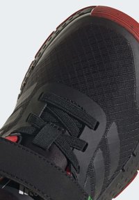 Svart atletisk sko med övre del i texturerad tyg, svarta snörningar, röd hälaccent och gummisula med greppmönster.