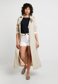 Beige lang trenchcoat med krage, knapp foran og rullede ermer, brukt over en svart topp, kombinert med hvite shorts og mønstrede sandaler.