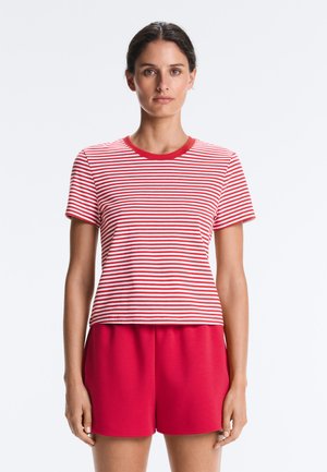 Vrouw met donker haar in een rood-wit gestreept kortemouwen T-shirt en effen rode korte broek, staand tegen een effen witte achtergrond.