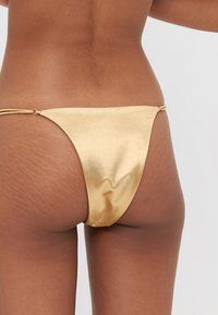 Zeltīta metāla bikini apakšdaļa ar gludu faktūru, minimālu pārklājumu un sasietām malām, kas aprīkotas ar mazām metāla gredzenēm regulēšanai.