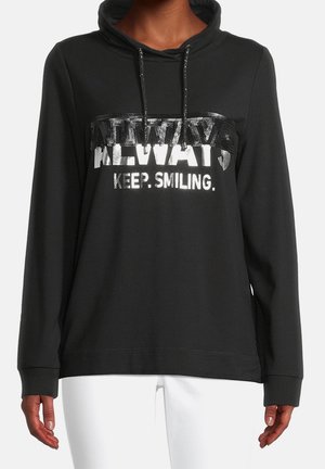 Sweat à capuche noir avec le texte en sequins « ALWAYS » et le texte argenté « KEEP. SMILING. » porté par une personne en pantalon blanc, mains détendues le long du corps.