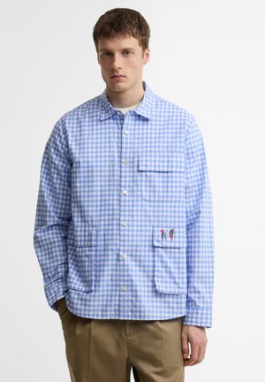 Fiatal férfi világoskék és fehér kockás gombos ingben, több zsebbel és apró hímzett részletekkel.