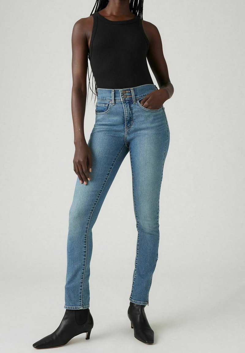 Jeans de tiro alto en azul con un corte ajustado, hechos de mezclilla. Combinados con una camiseta sin mangas negra y botas de tobillo negras. Tela suave con un leve desgastado.