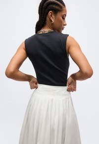 Paiga punutud juustega naine, kes kannab varrukateta musta topi ja valget volditud seelikut, reguleerib vöökohalt tagantpoolt ära.