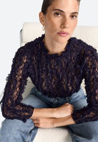 Top in pizzo blu navy con motivo floreale testurizzato, maniche lunghe e scollo a volant, abbinato a jeans in denim blu chiaro.