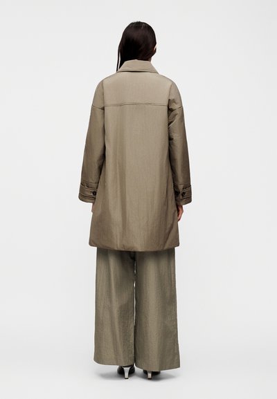 Γυναίκα που στέκεται με την πλάτη στην κάμερα, φορώντας χαλαρό μπεζ μακρύ παλτό, φαρδιά παντελόνια και μαύρες γόβες με φόντο έναν απλό λευκό τοίχο.