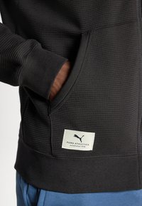 Hanorac negru cu textură, cu fermoar, cu manșete și tiv din material cu nervuri. Include un eticheta PUMA Athletics și buzunare laterale. Pantaloni albaștri parțial vizibili.