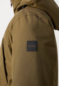 Gros plan d'une manche de veste marron avec un patch carré noir portant la mention BOSS sur le haut du bras.