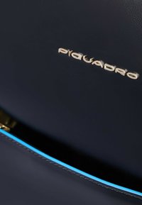 Bolso de cuero azul marino con logotipo dorado "Piquadro" y costuras de acento en azul. Presenta una textura suave y un diseño limpio y moderno.