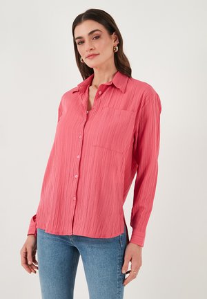 Pink long-sleeve button-up shirt with textured fabric, a chest pocket, and a curved hem, paired with blue jeans.