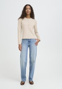 Bež dolg rokav s gumbi, kombiniran s svetlo modrimi kavbojkami z ravnim krojem. Model stoji na belem ozadju.