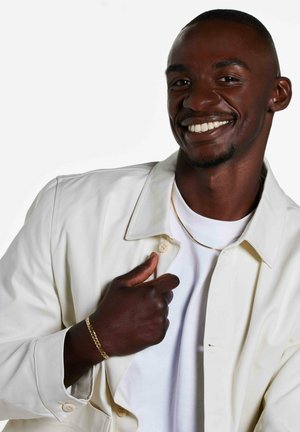 Homme souriant portant une veste blanche et un t-shirt, tenant le col de la veste, avec une chaîne en or et un bracelet sur fond blanc.