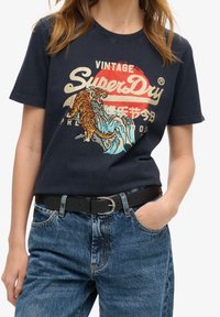 Mörkblå bomullströja med en grafisk design som har en tiger och vågor. Korta ärmar och rund halsringning, matchad med högmidjade jeans.