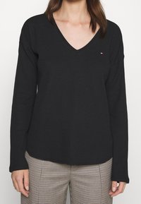 Czarna bluzka z długim rękawem i dekoltem w serek, wykonana z teksturowanej tkaniny, z małym logo na piersi. W zestawie z spodniami w kratę.