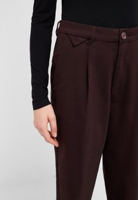 Burgundy trousers with a textured finish, featuring a button closure, side pockets, and pleated design. Hand slightly visible on waistband.