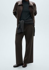 Brązowa skórzana kurtka na ciemnoszarym swetrze, z szarym swetrem związanym w talii. Luźne, szerokie brązowe spodnie oraz ciemne buty dopełniają strój.