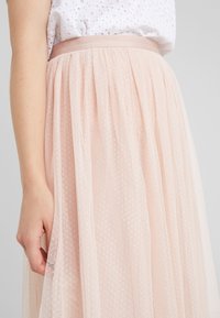 Mujer vistiendo una blusa blanca texturada y una falda de tul rosa claro de cintura alta con un patrón de pequeños puntos, con una mano descansando a un lado.