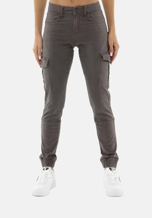Personne portant un pantalon cargo gris foncé avec des poignets élastiques et des baskets blanches, debout devant un fond clair uni.
