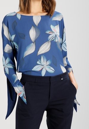 Blaues Blumenshirt mit langen Ärmeln und breiten Bündchen, aus einem leichten Stoff gefertigt. Auffällige Blumen- und Blattmuster in Pastellfarben.
