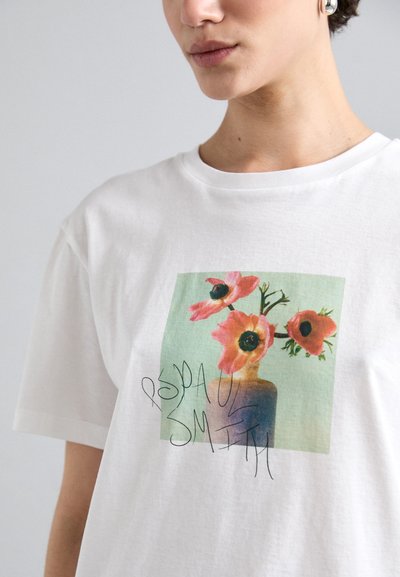 Hvit bomulls T-skjorte med et blomsterdesign som viser rosa blomster i en vase, akkompagnert av grønne og svarte detaljer, vist på forsiden.