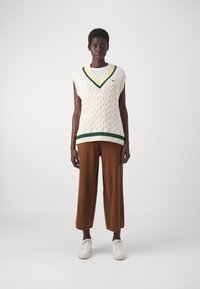 Pull sans manches en tricot crème avec un col en V, orné d'une bordure verte et jaune. Associé à un pantalon large marron et des baskets blanches.