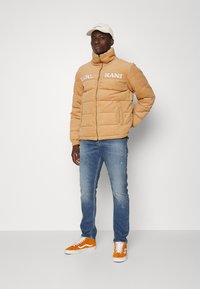 Brun pufferjakke med høy krage og logo-detaljer, kombinert med blå jeans og oransje joggesko. Modellen står foran en ensfarget bakgrunn.
