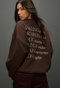 Barna kapucnis pulóver, bő szabással, fehér hímzett szöveggel a hátsó részen. A szöveg különböző stílusokban idézze a szezonális mondatokat.